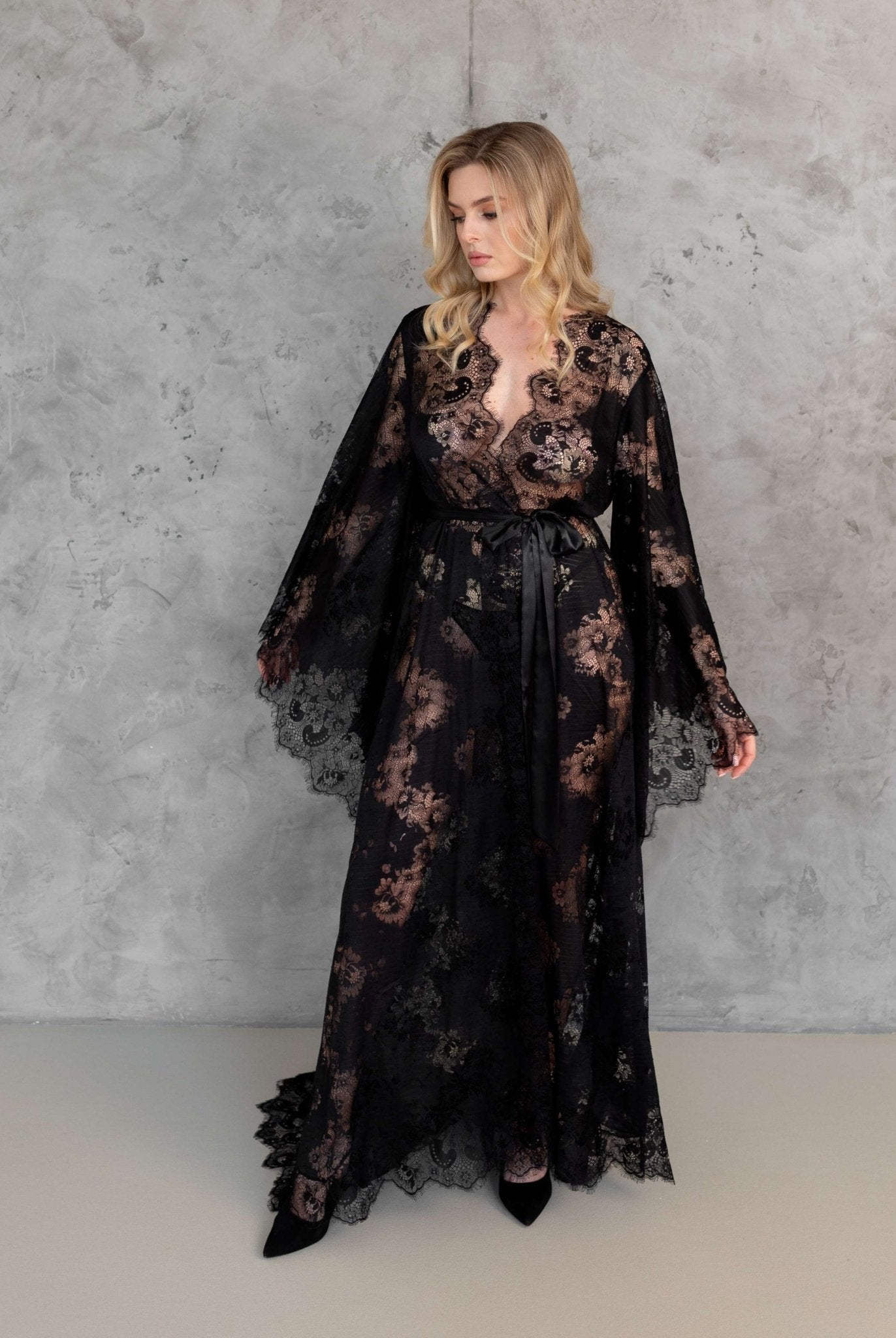 Model in sheer black lace robe with flared sleeves, posing in front of textured grey wall – KÂfemme