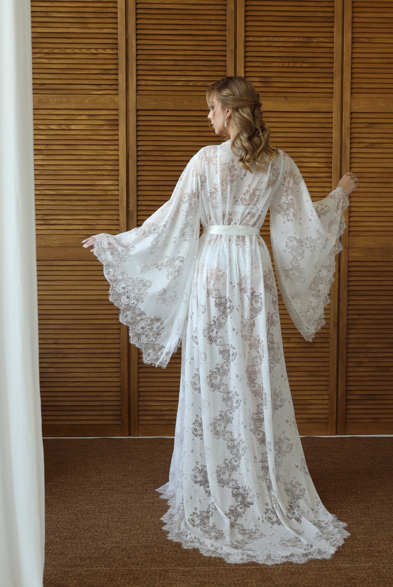Back view of white lace robe with sweeping train, posed by wooden backdrop – KÂfemme