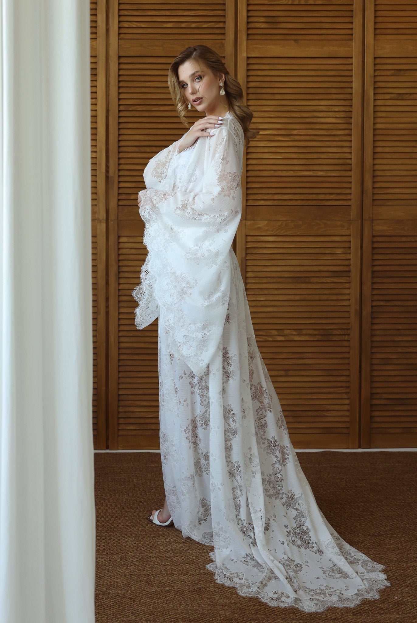 Model in sheer white lace robe with flared sleeves, standing against wooden background – KÂfemme