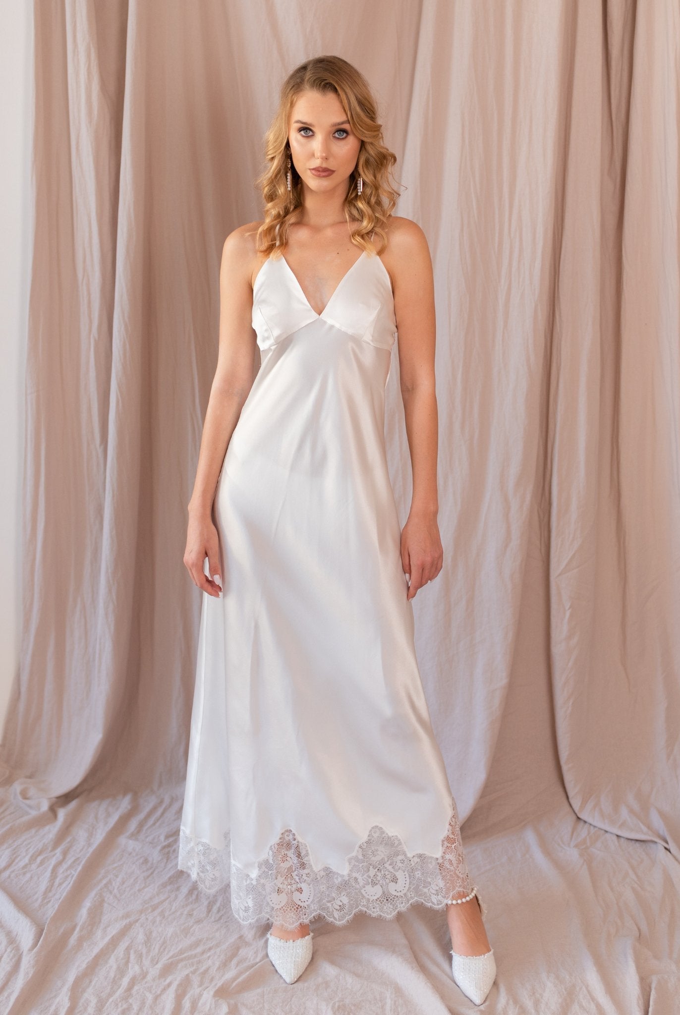 Woman standing in satin white nightgown with lace trim, light pink curtain background