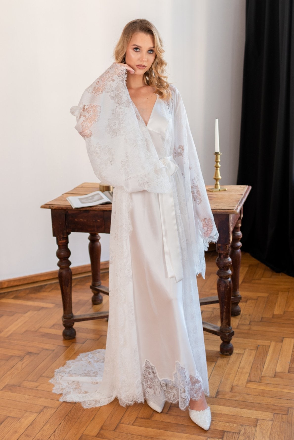 Woman standing in a white lace bridal robe and silk nightgown in a wood-floored room