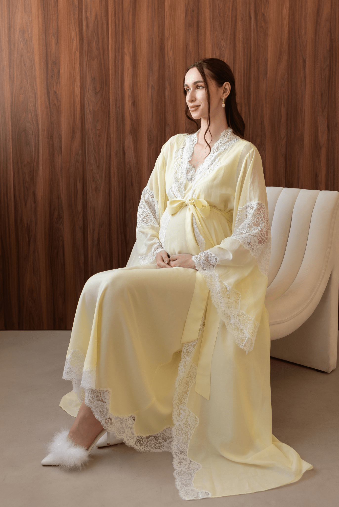 Pregnant woman sitting on a white chair, gently holding her belly in a comfortable yellow cotton lace ensemble.