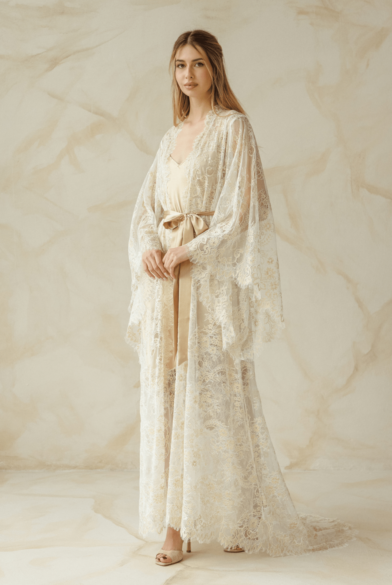 Side view of model gently draping long lace robe in soft beige tones