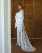 Model in sheer white lace robe with flared sleeves, standing against wooden background – KÂfemme