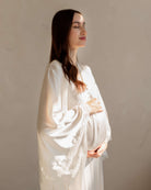 Gentle waist-up shot in a white set with sunlight, softly embracing a baby bump for a tender look.